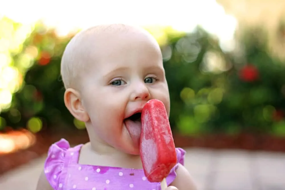 A cute 10 month old baby girl is eating a srawberry frozen fruit popsicle outside on a summer day.