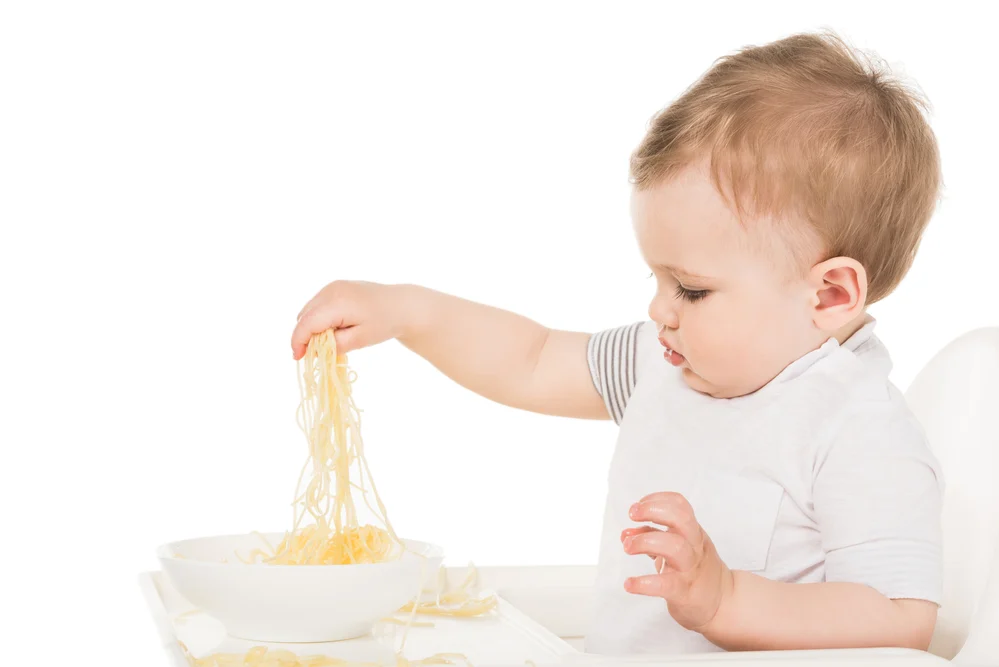 toddler sitting at high chair grabbing a handful of speghetti
