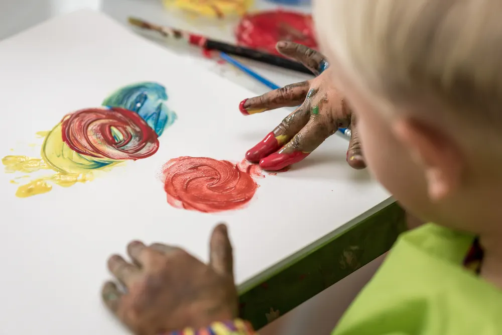 Over the shoulder view of a little boy having fun painting with fingers with colourful hand paints.