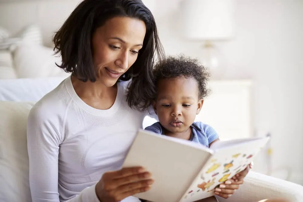 Close up of mixed race young adult mother sitting in an armchair reading a book with her two year old son, close up