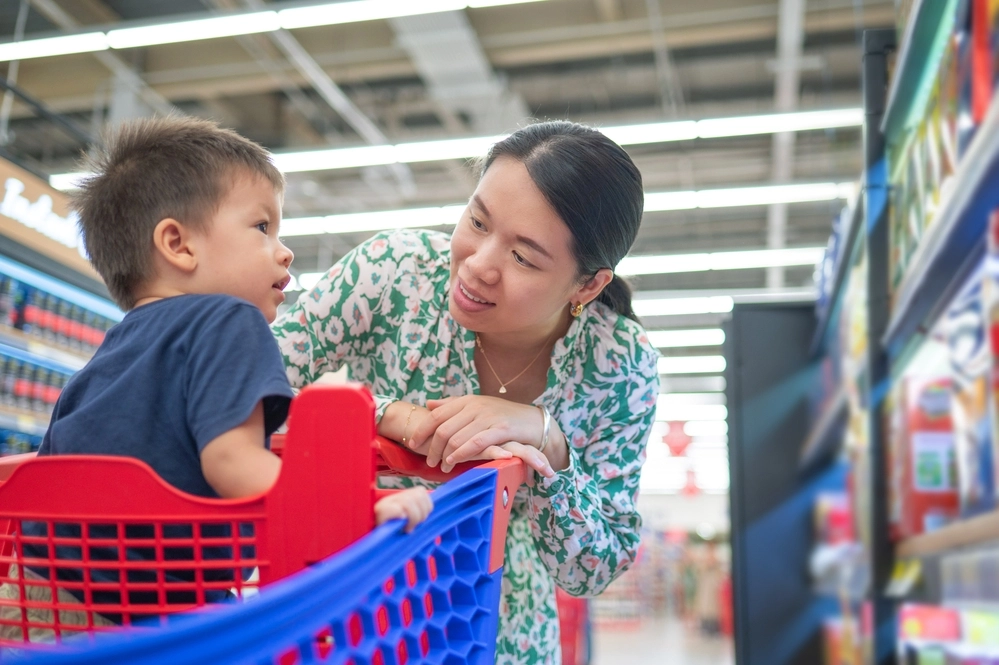 Young asian mom in green floral dress and her little multiracial boy in shopping trolley buying groceries in supermarket